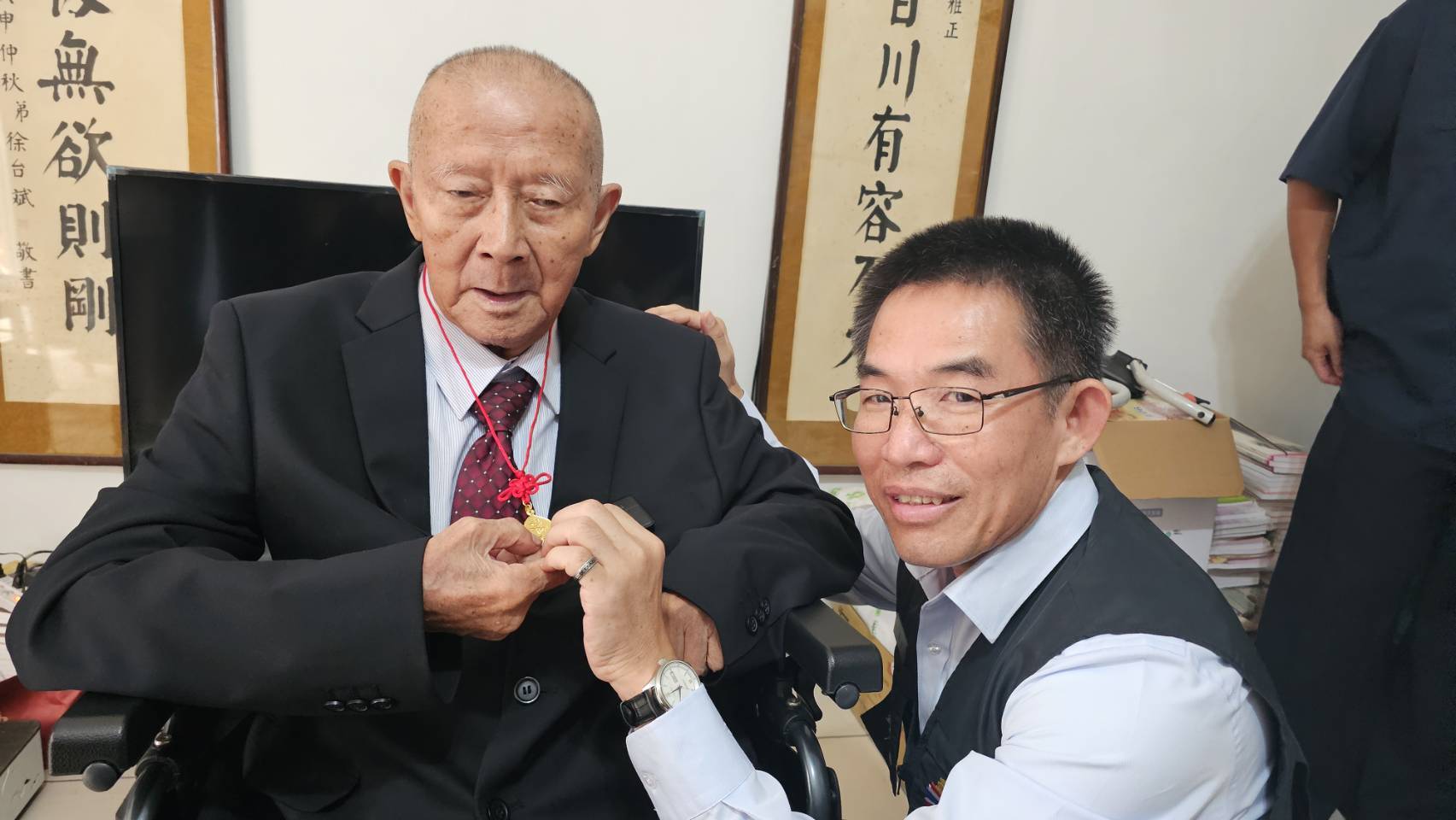 臺北市榮民服務處處長黃信仁前往鍾爺爺住家祝壽，代表輔導會主委嚴德發致贈金壽桃項鍊，並親自為鍾爺爺細心佩掛。（臺北市榮服處提供）
